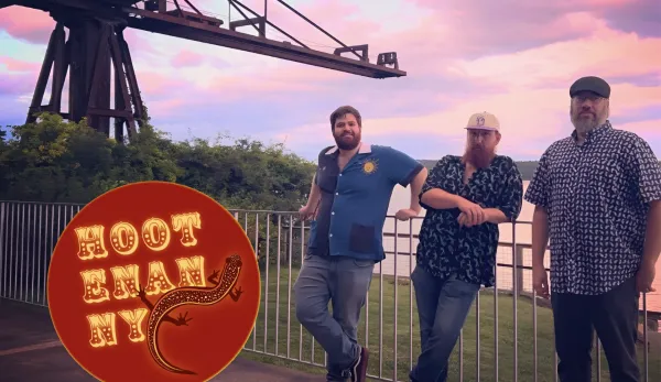 Three men pose in front of a railing and an industrial structure, with a circular "HOOTENANNY" logo,